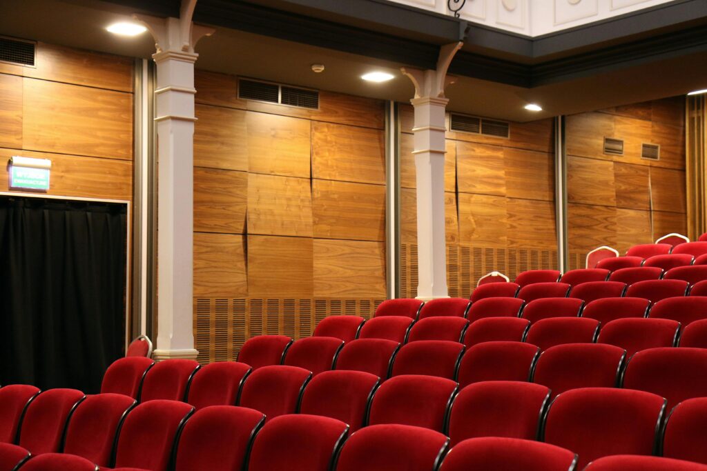Blog Interior of an empty auditorium with red seats and wooden panels, lit overhead.